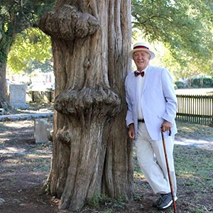 Man in white suit and straw hat by a tree in the burying ground